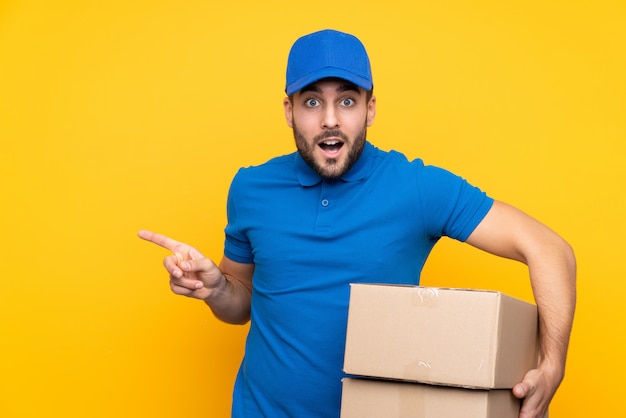 Delivery man over isolated yellow wall surprised and pointing finger to the side