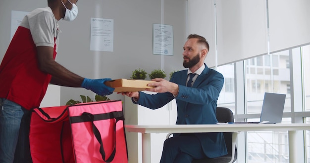 Delivery man in medical face mask and gloves giving pizza boxes tamale customer at office