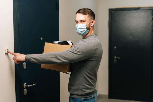 Delivery man in medical mask holding cardboard box and\
contactless payment pos wireless terminal for card paying ringing\
doorbell