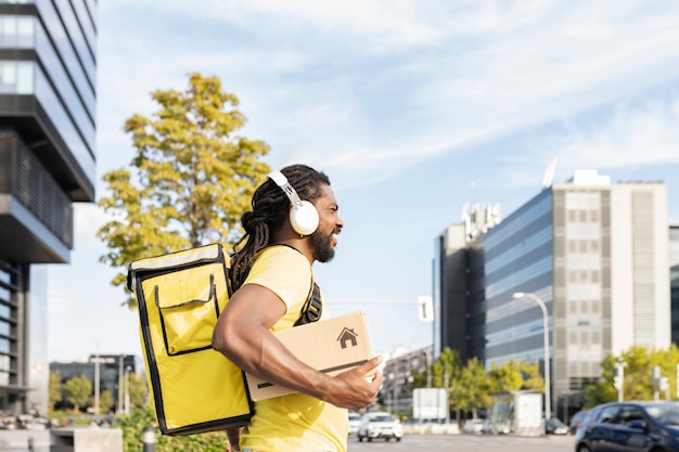 Delivery man with backpack and a box in his hand, listening to music in the city