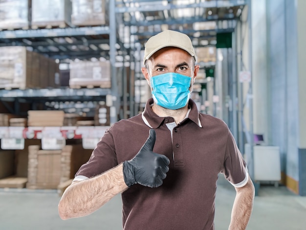 Delivery man with uniform and protective mask for covid-19 inside a warehouse. Safe shipping concept.