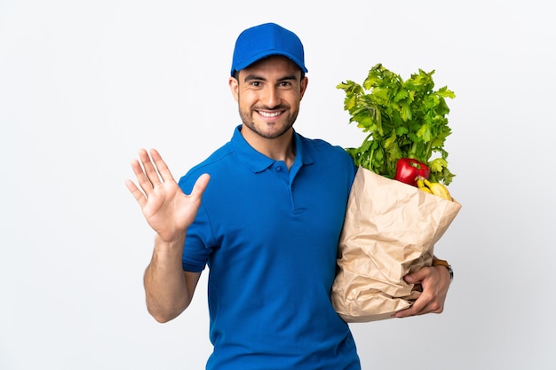 Delivery man with vegetables isolated on white wall saluting with hand with happy expression