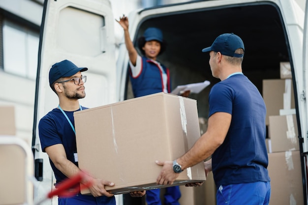 Delivery men cooperating while loading cardboard boxes in a mini van Their female colleagues is gesturing from a van