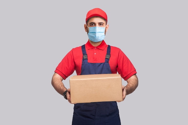 Consegna in quarantena. ritratto di giovane uomo con maschera medica chirurgica in uniforme blu e maglietta rossa in piedi e tenendo la scatola di cartone su sfondo grigio. indoor, girato in studio, isolato,