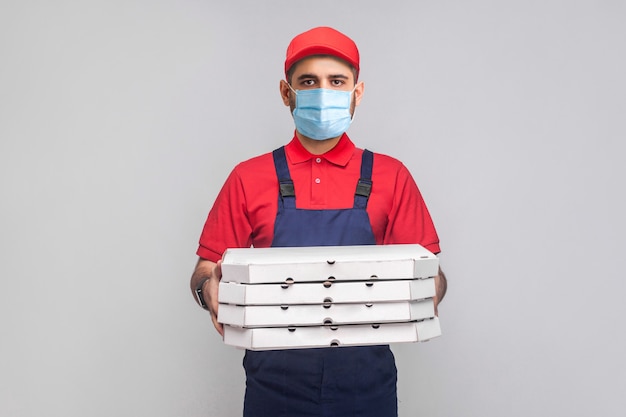 Delivery on quarantine. Young man with surgical medical mask in blue uniform and red t-shirt standing and holding stack of cardboard pizza boxes on grey background. Indoor, studio shot, isolated,