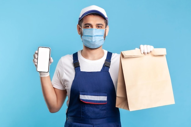 Delivery service mobile app Courier man in uniform  mask showing parcel and cellphone with blank display mock up for express shipping advertise online takeaway food order indoor shot isolated