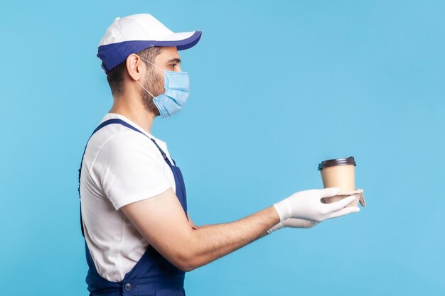 Delivery service Side view friendly happy courier man in overalls mask offering coffee wearing safety gloves giving drinks in disposable cups and smiling studio shot isolated on blue background