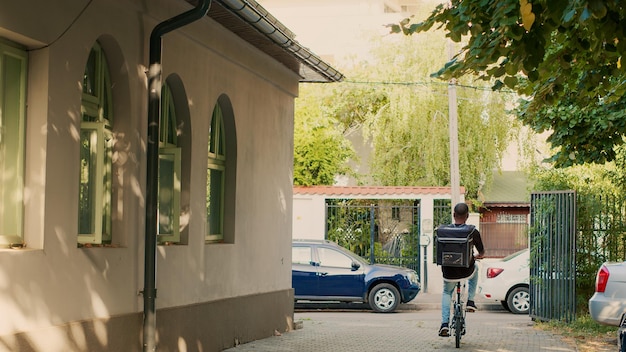 Delivery service worker with thermal backpack riding bicycle, delivering fastfood takeaway order at front door. Male courier using bike to give restaurant food and lunch meal. Paperbag package.