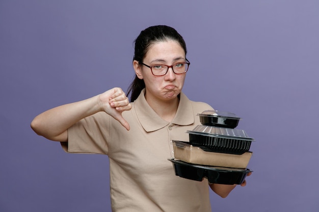 delivery woman employee in blank polo shirt uniform wearing glasses holding food containers looking at camera being displeased showing thumb down standing over blue background