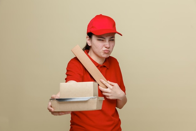 Delivery woman employee in red cap and blank tshirt uniform holding food boxes looking at camera being displeased making wry mouth standing over brown background