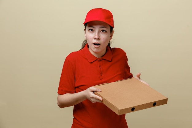 Dipendente donna di consegna in berretto rosso e maglietta bianca uniforme che tiene scatola per pizza guardando la fotocamera stupito e sorpreso in piedi su sfondo marrone