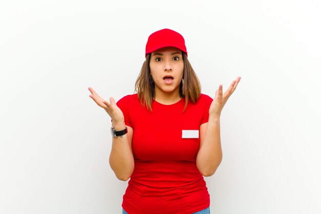 Delivery woman feeling happy, excited, surprised or shocked, smiling and astonished at something unbelievable against white wall