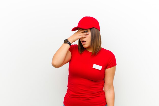 Delivery woman looking shocked, scared or terrified, covering face with hand and peeking between fingers on white