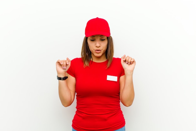 Delivery woman with open mouth pointing downwards with both hands, looking shocked, amazed and surprised against white