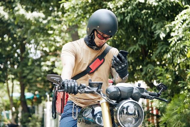 Photo deliveryman in helmet sitting on motorcycle and checking address of customer via application on smartphone