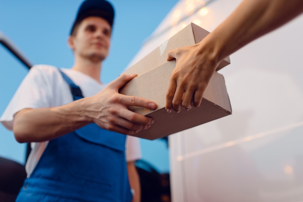 Deliveryman in uniform gives parcel to female recipient at the car, delivery service. Man holding cardboard package near the vehicle, male deliver and woman, courier or shipping job