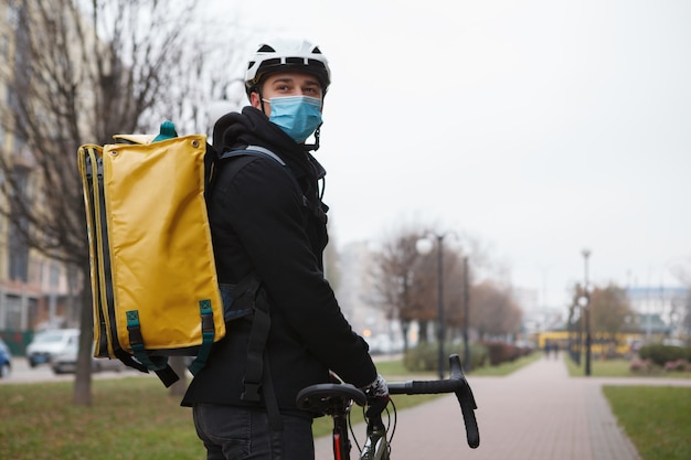 Fattorino che indossa maschera medica e zaino termico, guardandosi alle spalle mentre cammina con la sua bicicletta