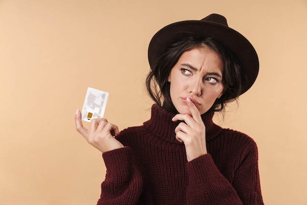 denken jonge brunette vrouw poseren geïsoleerd over beige muur muur met creditcard.