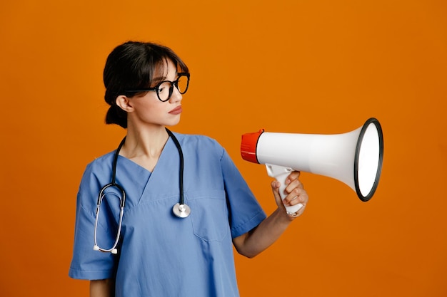 Denken met luidspreker jonge vrouwelijke arts dragen uniform fith stethoscoop geïsoleerd op oranje achtergrond