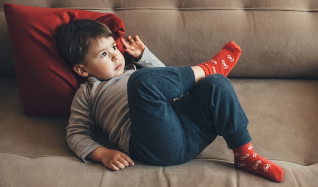 Denkende blanke jongen liggend op de bank met een rood kussen wegkijken in een zonnige dag