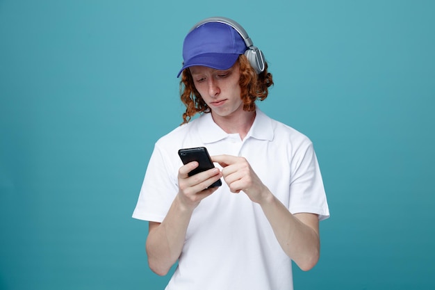 Denkende jonge knappe man in pet met koptelefoon die de telefoon vasthoudt en bekijkt die op een blauwe achtergrond wordt geïsoleerd