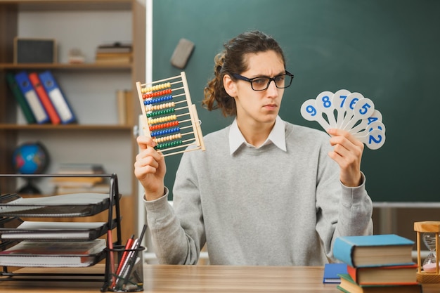 Foto denkende jonge mannelijke leraar met een bril die een telraam vasthoudt en bekijkt met nummerplezier aan een bureau zitten met schoolhulpmiddelen in de klas