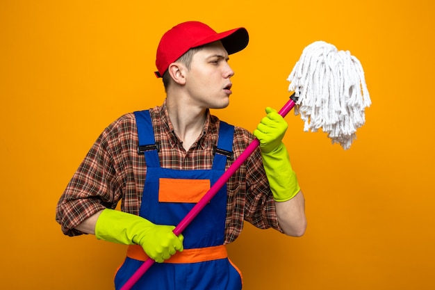 Denkende jonge schoonmaakster die uniform en pet draagt met handschoenen die de dweil vasthouden en bekijken