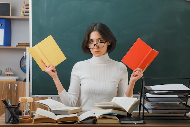 Denkende jonge vrouwelijke leraar met een bril die boeken vasthoudt die aan een bureau zitten met schoolhulpmiddelen in de klas