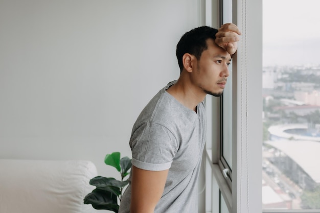 Denkende serieuze eenzame aziatische man staat leunend op het raam in zijn appartement