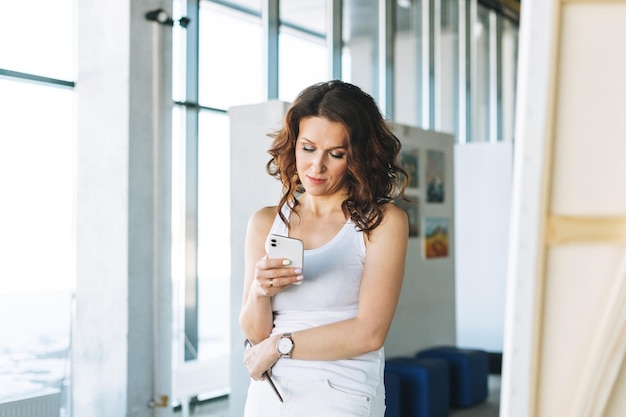 Denkende vrouw kunstenaar in witte kleding met kwast met behulp van mobiel voor ezel in kunstgalerie