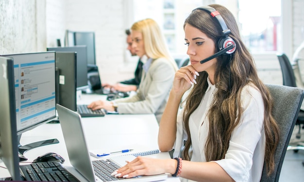 Denkende zakenvrouw die op een headset praat terwijl ze op een laptop werkt in het kantoor