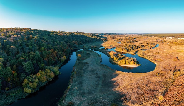 Dense green forest in shade and broad grassy meadow near long curving river branches under cloudy sky at sunset panorama view