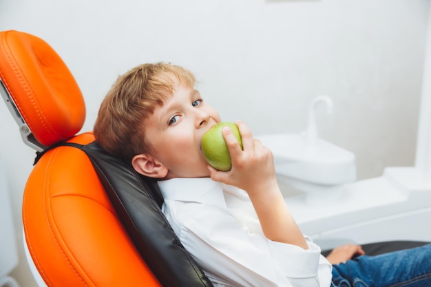 Стоматологическая клиника Уход за зубами Маленький мальчик держит яблоко, сидя в стоматологическом кресле