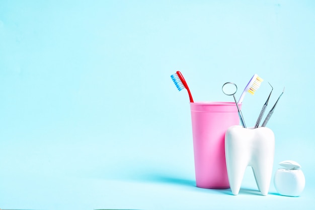 Dental mirror with explorer probes in healthy white tooth model near dental floss and toothbrushes in pink glass on light blue background