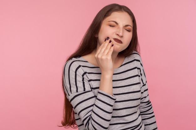 Problemi dentali ritratto di donna malata infelice in felpa a righe che tocca la guancia dolorante accigliata soffrendo di mal di denti cavità o smalto sensibile indoor studio girato isolato su sfondo rosa