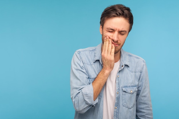 Dental problems. Portrait of unhealthy man in denim shirt pressing sore cheek, suffering acute toothache, periodontal disease, cavities or jaw pain. indoor studio shot isolated on blue background