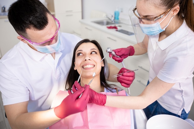 Foto cure odontoiatriche durante la quarantena del coronavirus. sorridente giovane donna in una poltrona del dentista. dentista e assistente in maschere e guanti protettivi controllano i denti del paziente