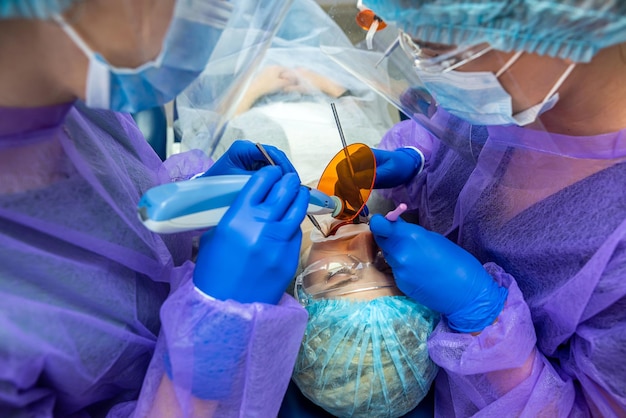 Dentist and assistant in blue latex gloves and masks fill the\
teeth of his patient in goggles