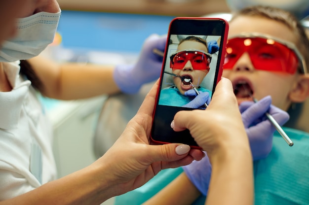 歯科医院での歯の治療中に歯科用椅子に座っている少年の写真撮影をしている歯科助手。スマートフォンに焦点を当てる