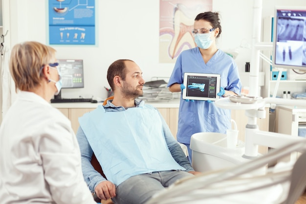 Dentist assistant showing teeth radiography on tablet pc computer screen to sick patient man at dental clinic office