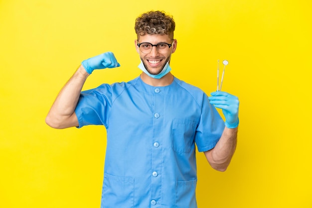 Dentist blonde man holding tools isolated on background doing strong gesture