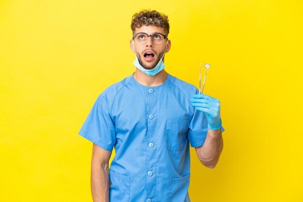 Dentista uomo biondo tenendo gli strumenti isolati su sfondo guardando in alto e con espressione sorpresa