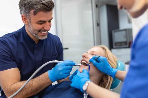 Dentist in blue uniform with assistant doing teeth procedure to patient female patient with turbine during treatment procedure in contemporary dental clinic