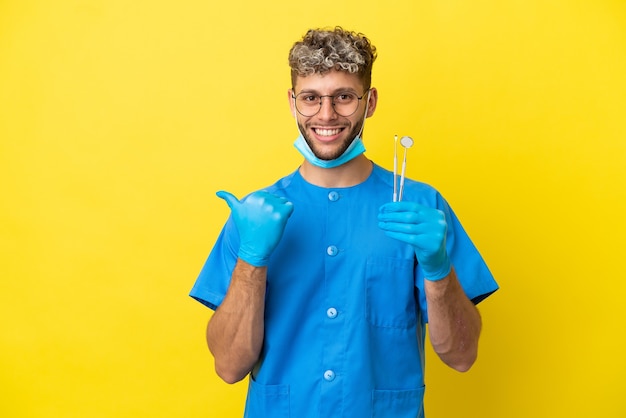 Dentist caucasian man holding tools isolated on yellow background pointing to the side to present a product