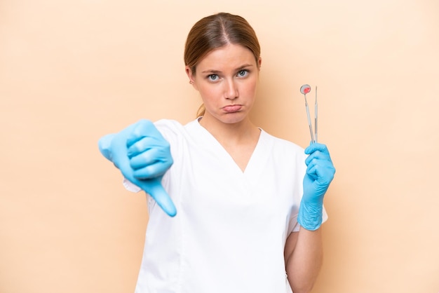 否定的な表情で親指を示すベージュの背景に隔離されたツールを保持している歯科医の白人女性