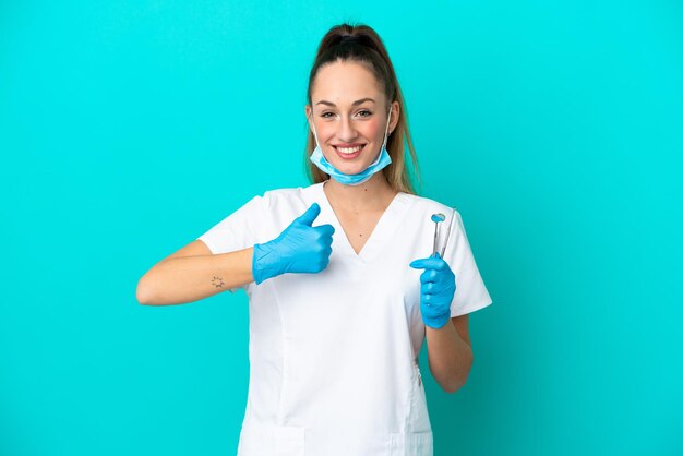 Стоматолог кавказская женщина, держащая инструменты, изолированные на синем фоне, показывая большой палец вверх жест