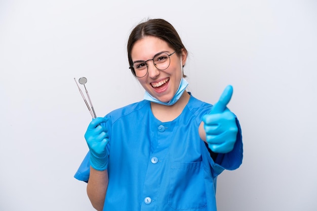 Стоматолог кавказская женщина держит инструменты на белом фоне с поднятыми большими пальцами, потому что произошло что-то хорошее