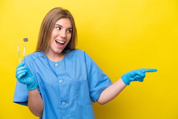 Dentist caucasian woman holding tools isolated on yellow background pointing finger to the side and presenting a product