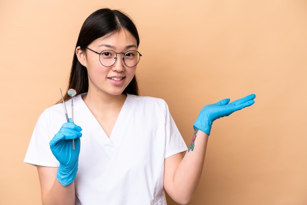 Dentist Chinese woman holding tools isolated on beige background extending hands to the side for inviting to come