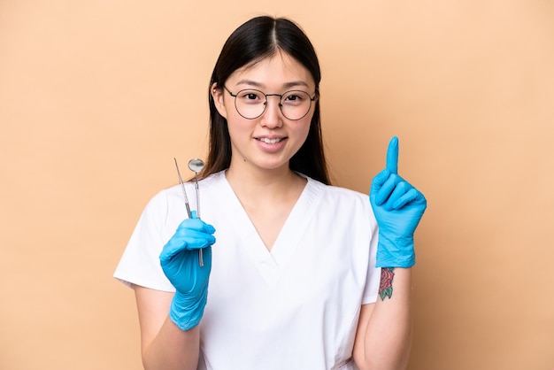 Dentist Chinese woman holding tools isolated on beige background showing and lifting a finger in sign of the best